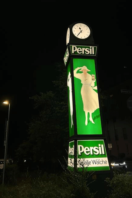 Vintage Litfaßsäule in grün mit alter Persil-Werbung und Frau in einem weißen Kleid. Oben auf der Säule ist eine Uhr angebracht.